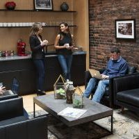 Three women and one man sitting in the atrium of the coworking space KNOWN. The man is working on a computer. Two women are talking to each other as they are each enjoying a coffee.